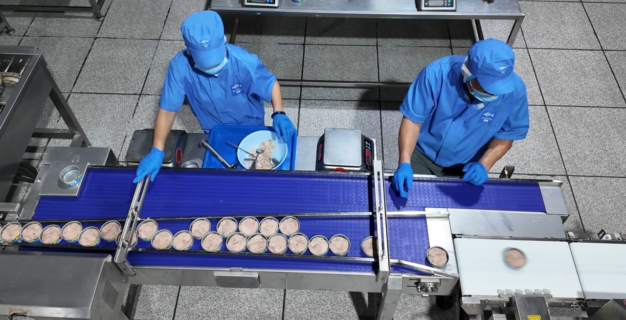Workers filling and weight-checking tuna cans on the line
